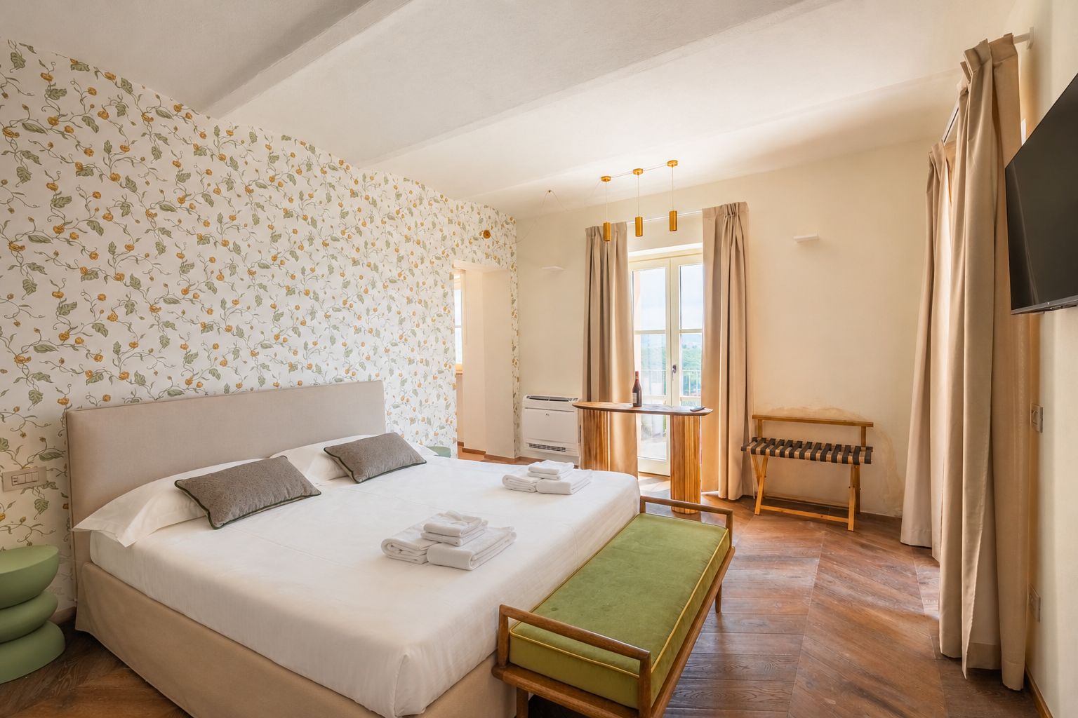 Quarto de hotel com cama, um banquinho, uma mesa com uma garrafa e uma televisão.