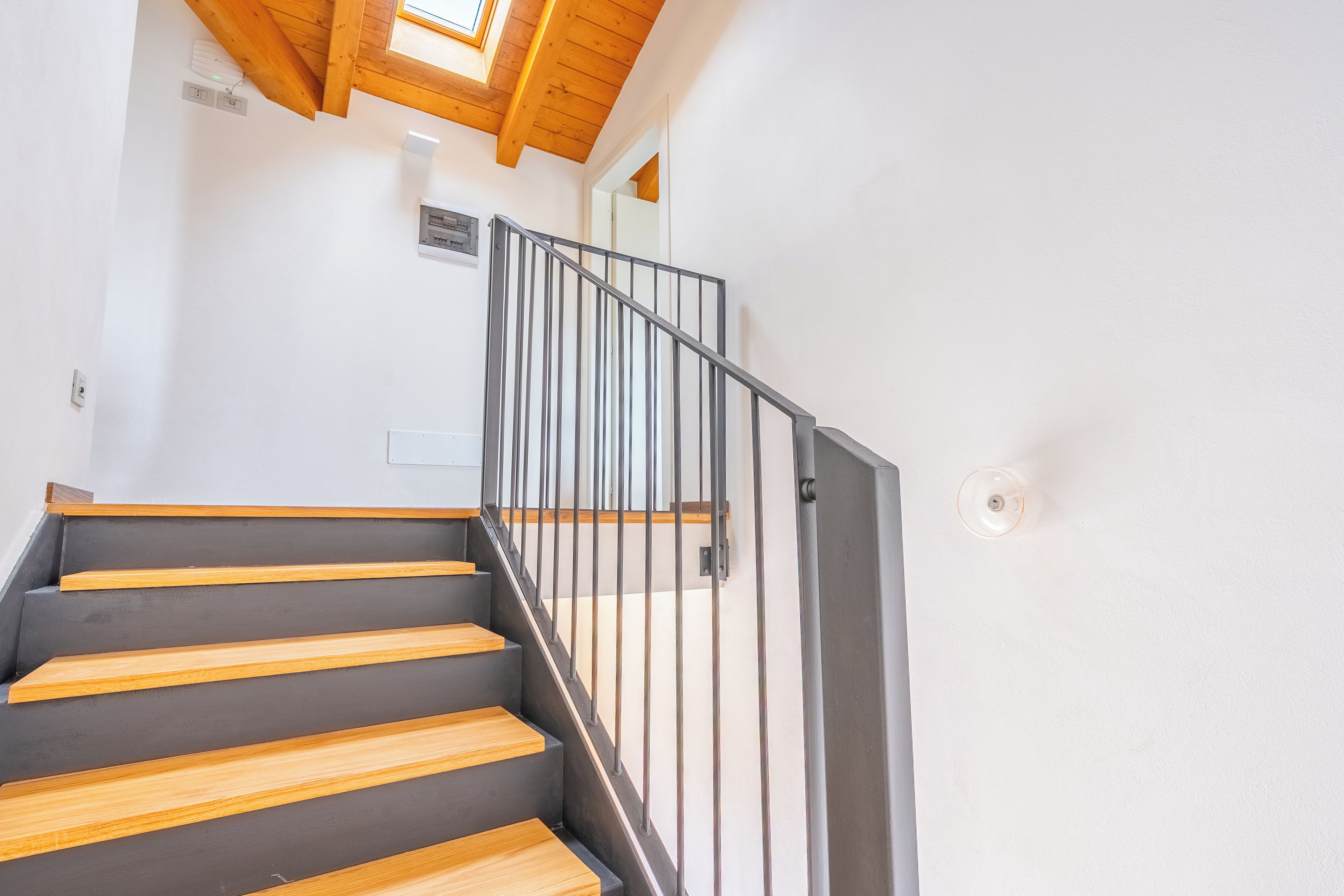 Un escalier avec des marches en bois et une rampe en métal, menant à une pièce dotée d’une lucarne.
