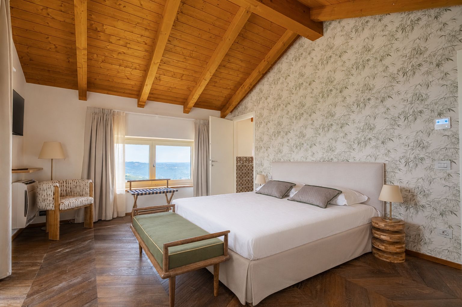 A bedroom with a large bed, wooden beams, a window with a view, a desk with a chair, and a patterned wall.