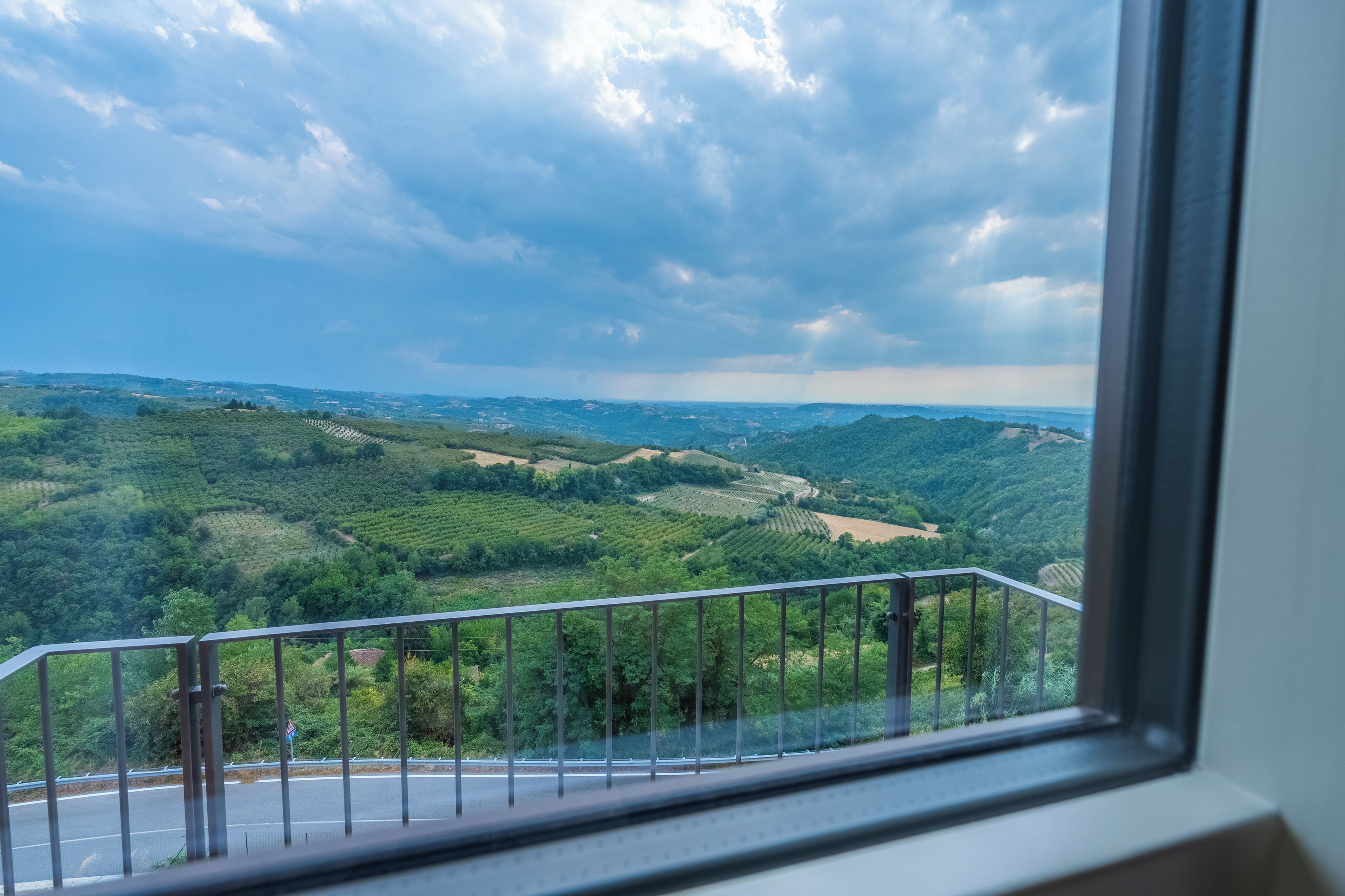 Una vista dalla finestra con ringhiera metallica, su un paesaggio con campi verdi e una strada.