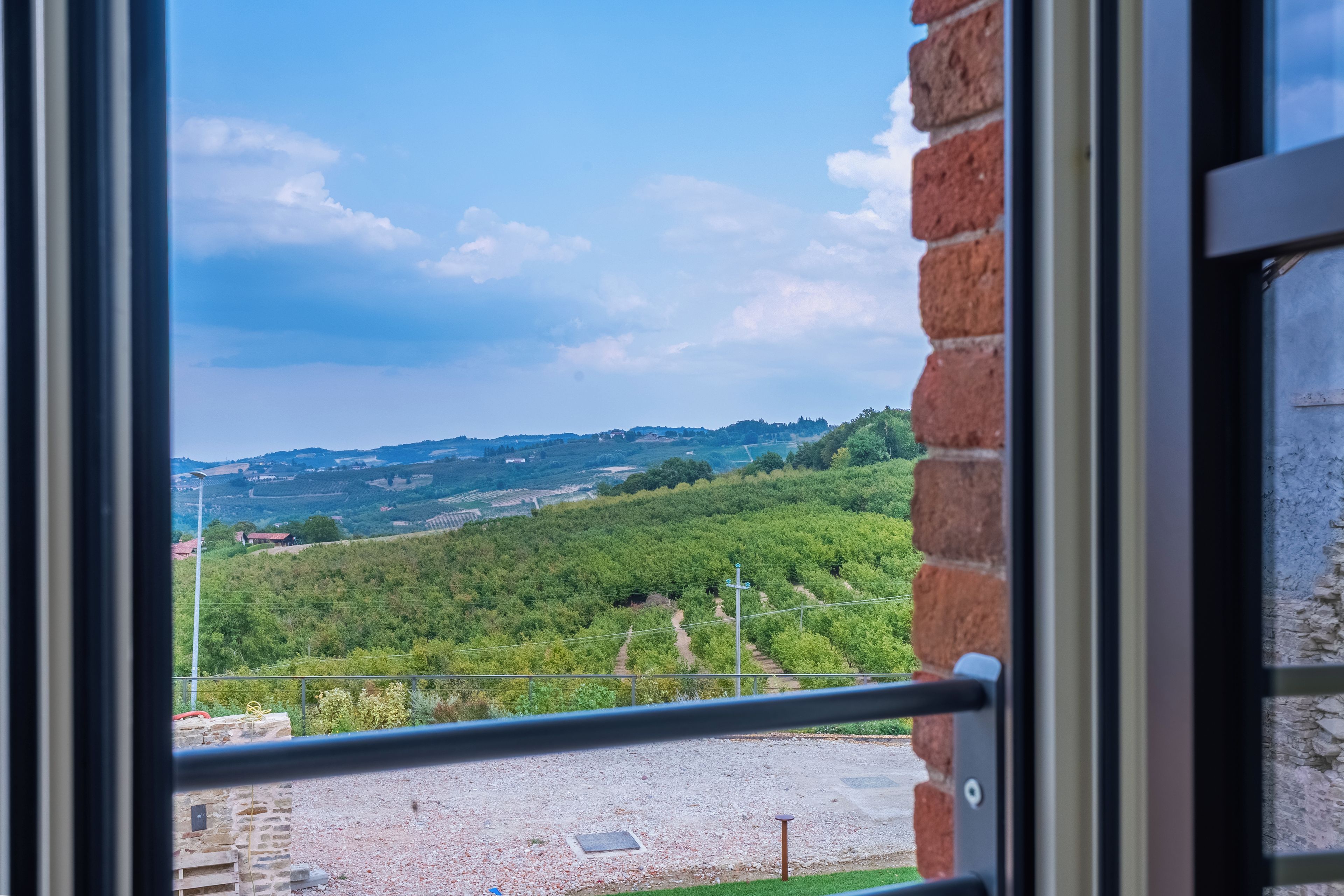 Une vue à travers une fenêtre, avec un mur de briques, un chemin de gravier et un paysage verdoyant, avec des arbres et des collines à l’horizon.
