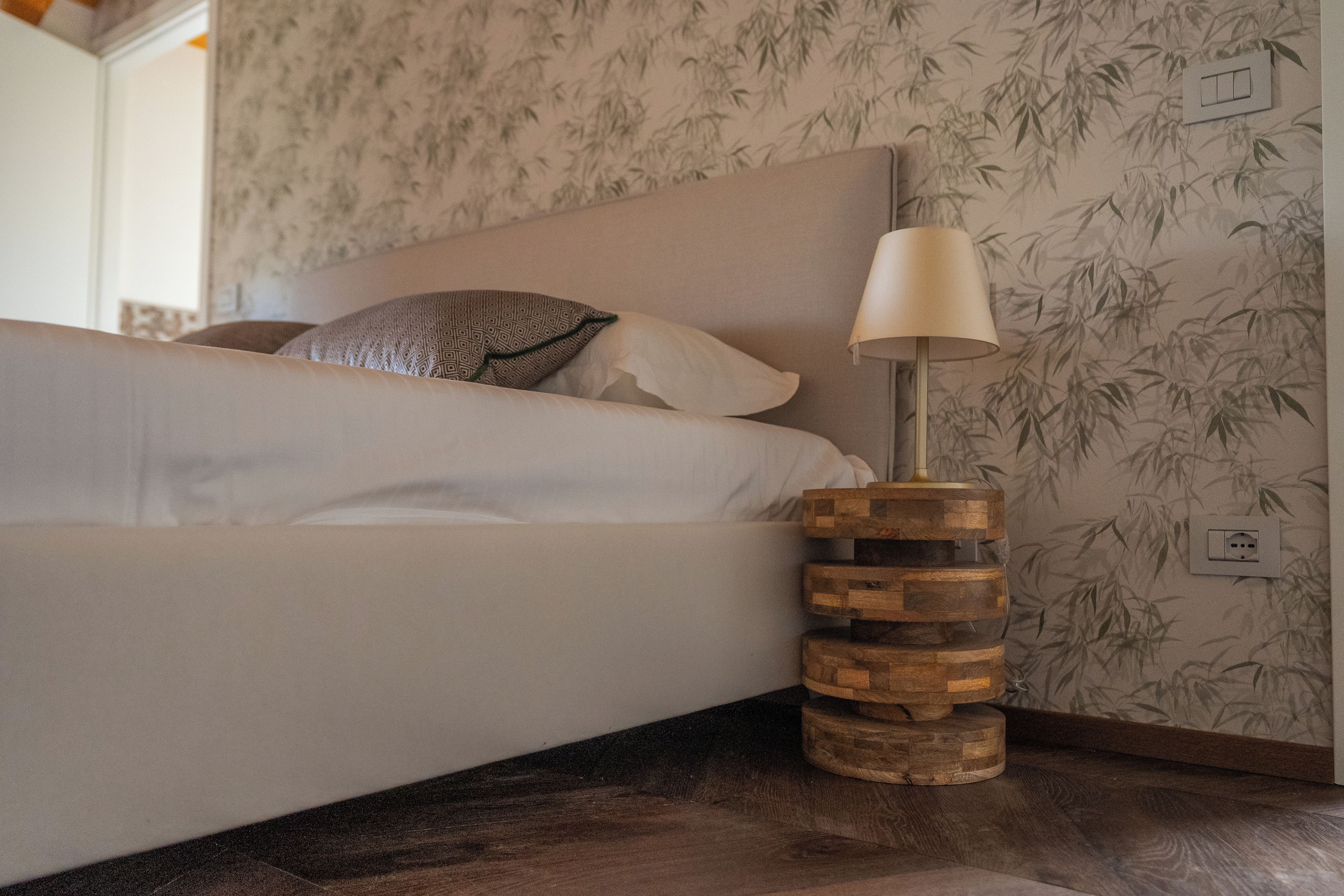 Un letto con un cuscino decorato, una lampada da comodino su un piedistallo in legno e una parete rivestita con carta da parati a motivi di bambù.