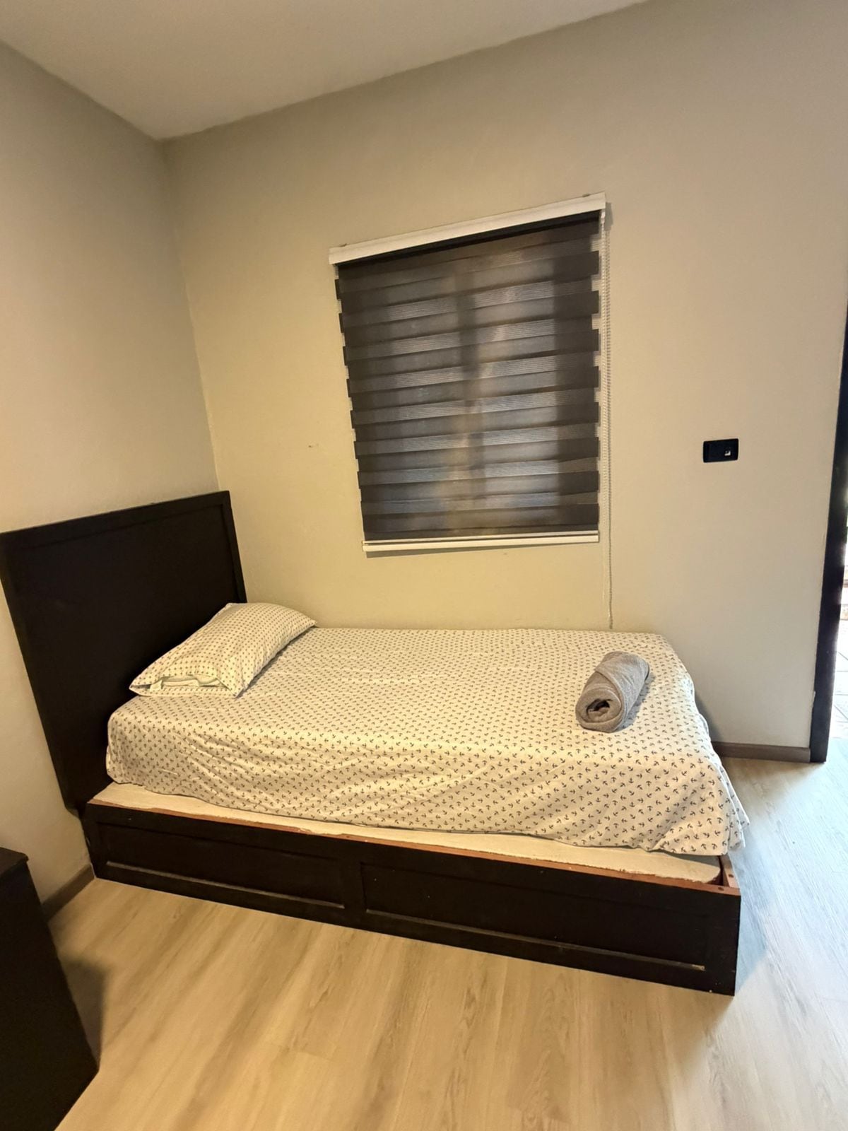 Una cama individual con cabecera, una ventana con persianas y un interruptor de luz en la pared.