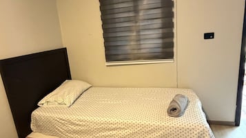 Una cama individual con cabecera, una ventana con persianas y un interruptor de luz en la pared.
