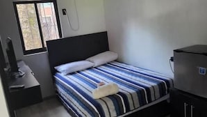 En enkeltseng med et stribet sengetæppe, et natbord med en lampe, et fjernsyn og et vindue med udsigt til bygninger.
