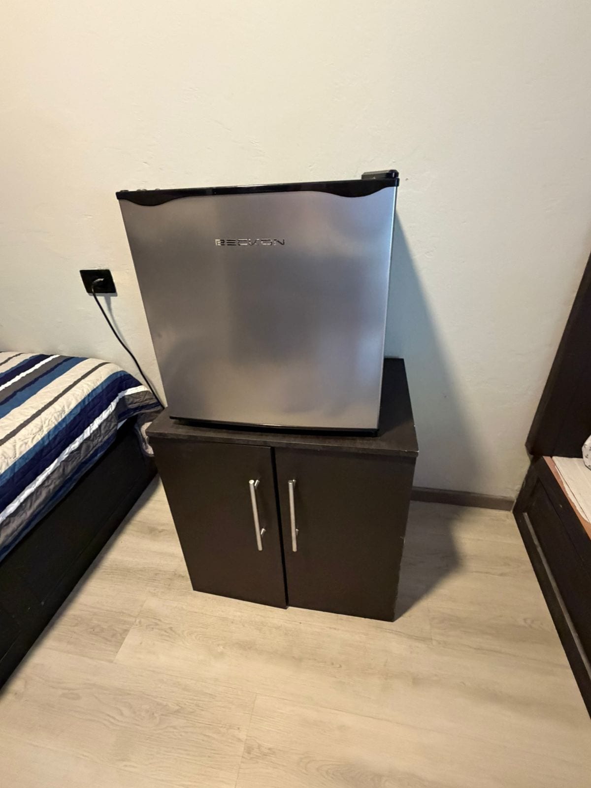 Un refrigerador compacto sobre un mueble bajo en un dormitorio con cama y mesita de noche.