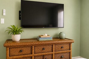 Un téléviseur fixé au mur, une console en osier dotée de tiroirs, une plante en pot et quelques livres.
