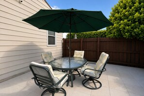 Une terrasse avec une table ronde en verre et quatre chaises, un parasol et une clôture en bois en arrière-plan.