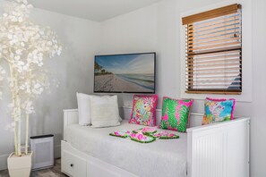 Ein Schlafzimmer mit einem Bett, einem an der Wand montierten Fernseher, einem Fenster mit Jalousien und einer Zierpflanze.