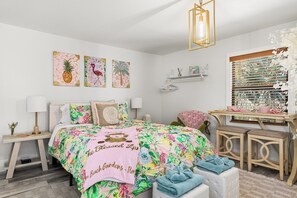 A bedroom with a floral bedspread, a bedside table with a lamp, a window with blinds, and wall art. - Palm Beach Pink Studio Farm Stay- Palm Tree & Alpaca / Llama Farm (Palm Beach Gardens)