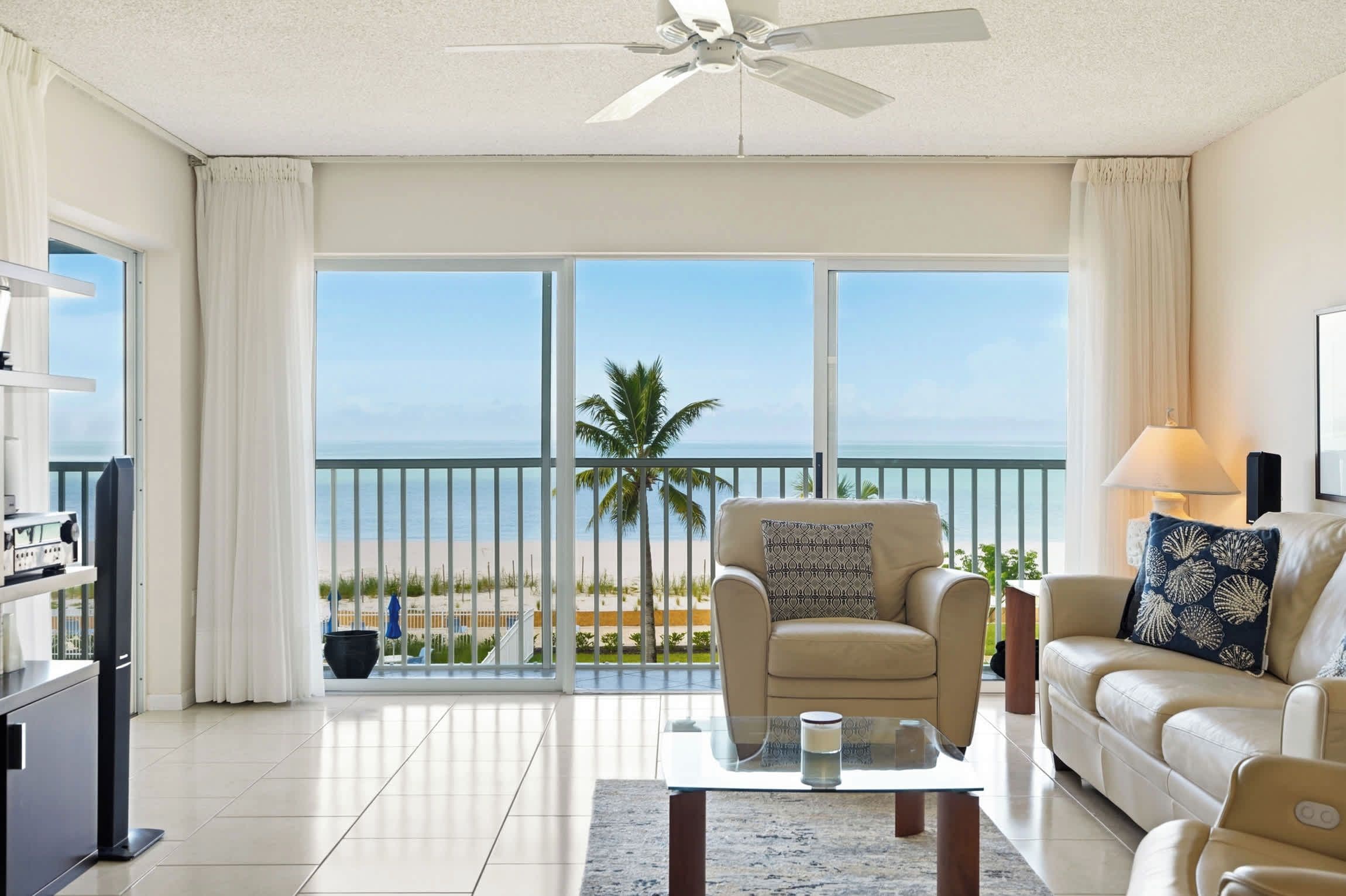 A living room with a view of the beach, a ceiling fan, and a sofa with decorative pillows.