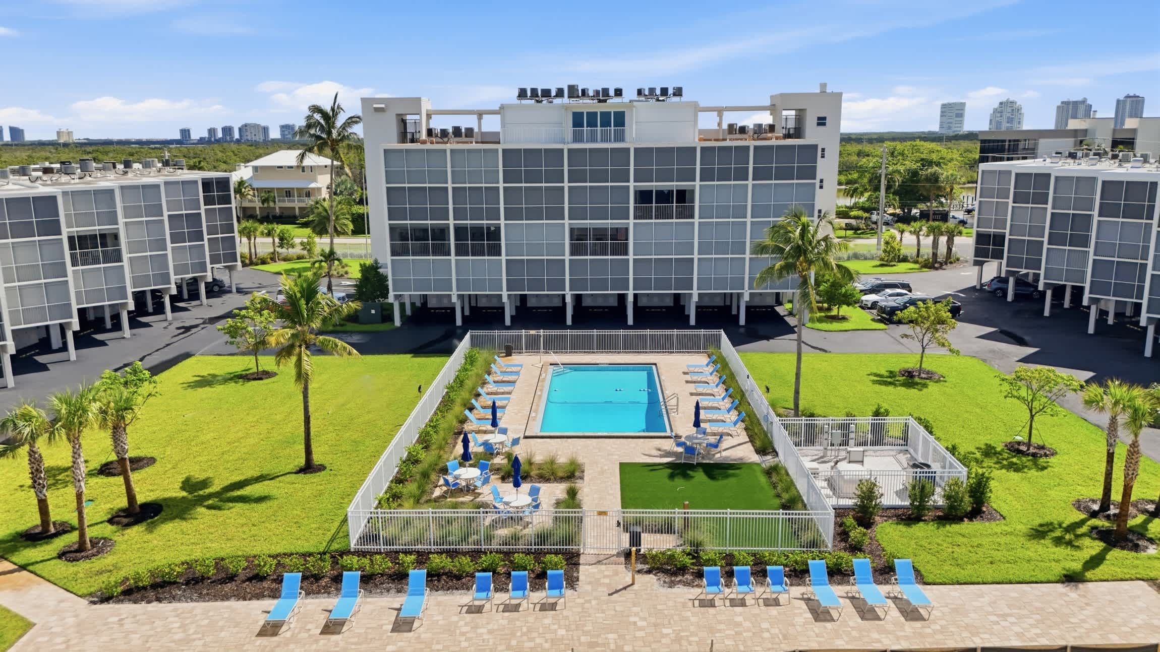 A modern building with a rooftop pool and lounge area, surrounded by greenery and palm trees.