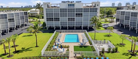 A modern building with a rooftop pool and lounge area, surrounded by greenery and palm trees.