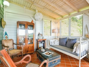 A room with a wooden ceiling, a white bed with blue pillows, a wicker chair, a cabinet with glass doors, and a fan.