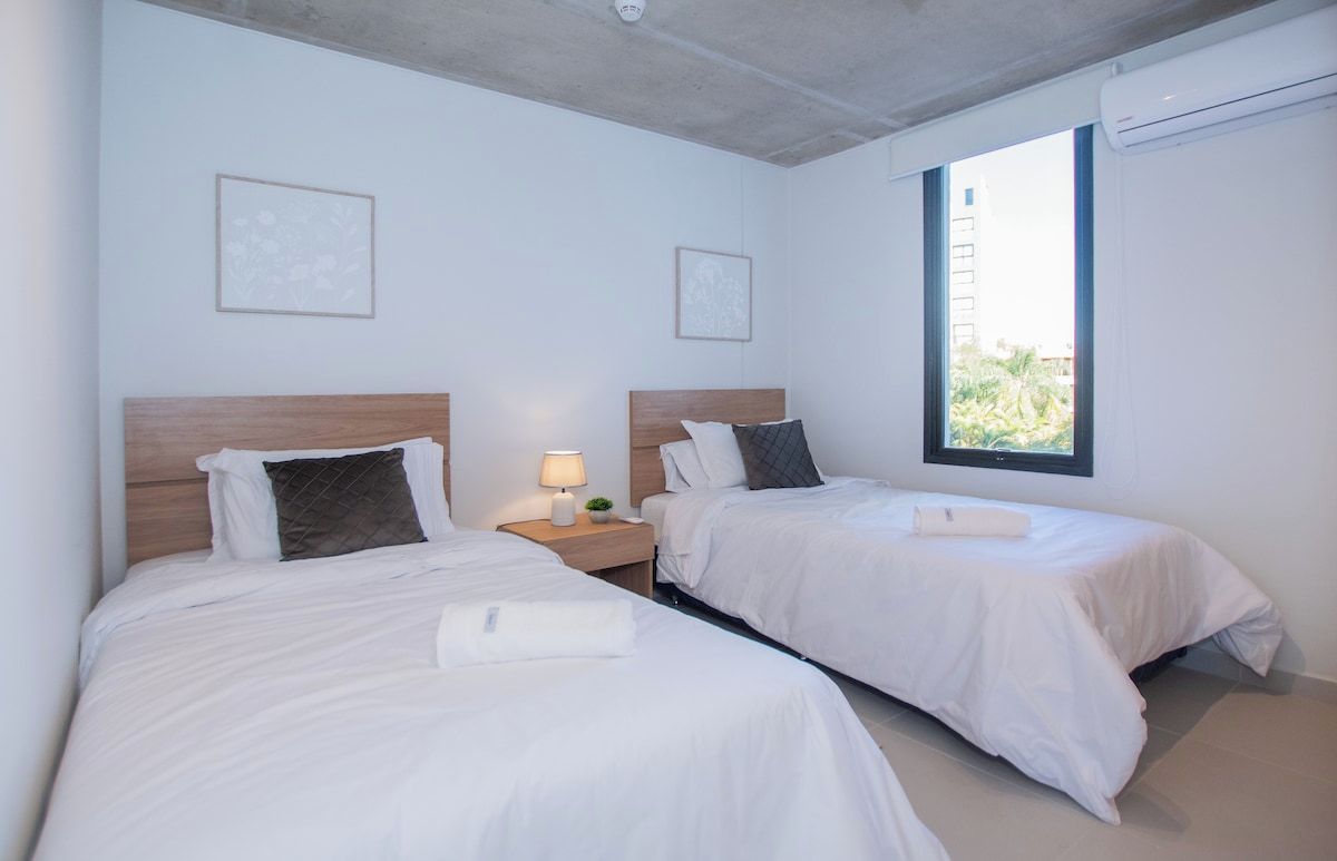 A hotel room with two beds, a nightstand with a lamp, and a window with a view.