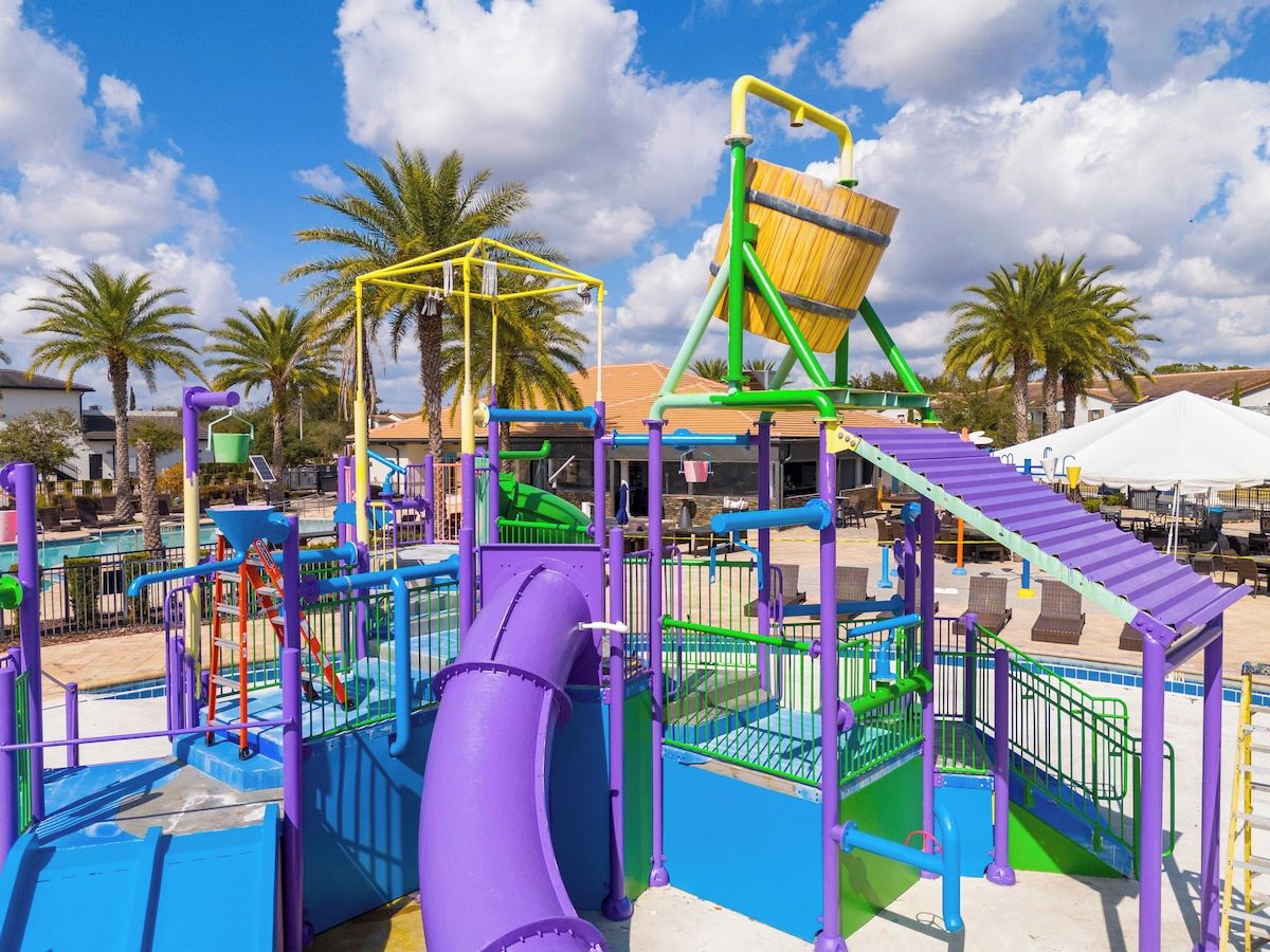 A playground with various play structures, including slides and climbing areas, surrounded by palm trees and a clear sky.