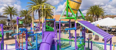 A playground with various play structures, including slides and climbing areas, surrounded by palm trees and a clear sky.