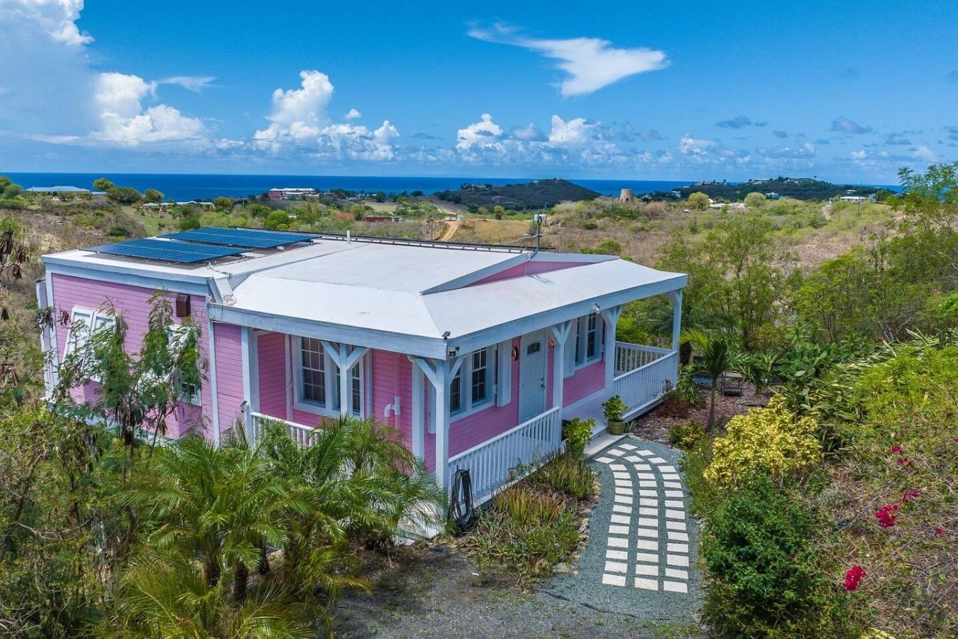 Une maison rose et blanche avec un panneau solaire sur le toit, entourée de verdure et offrant une vue dégagée sur l’océan.