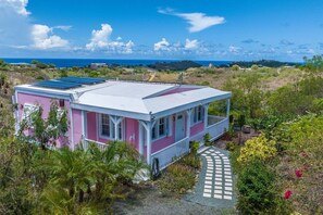 Una casa de color rosa i blanc amb un panell solar a la teulada, envoltada de vegetació i una vista clara del mar.