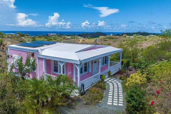 A pink and white house with a solar panel on the roof, surrounded by greenery and a clear view of the ocean.