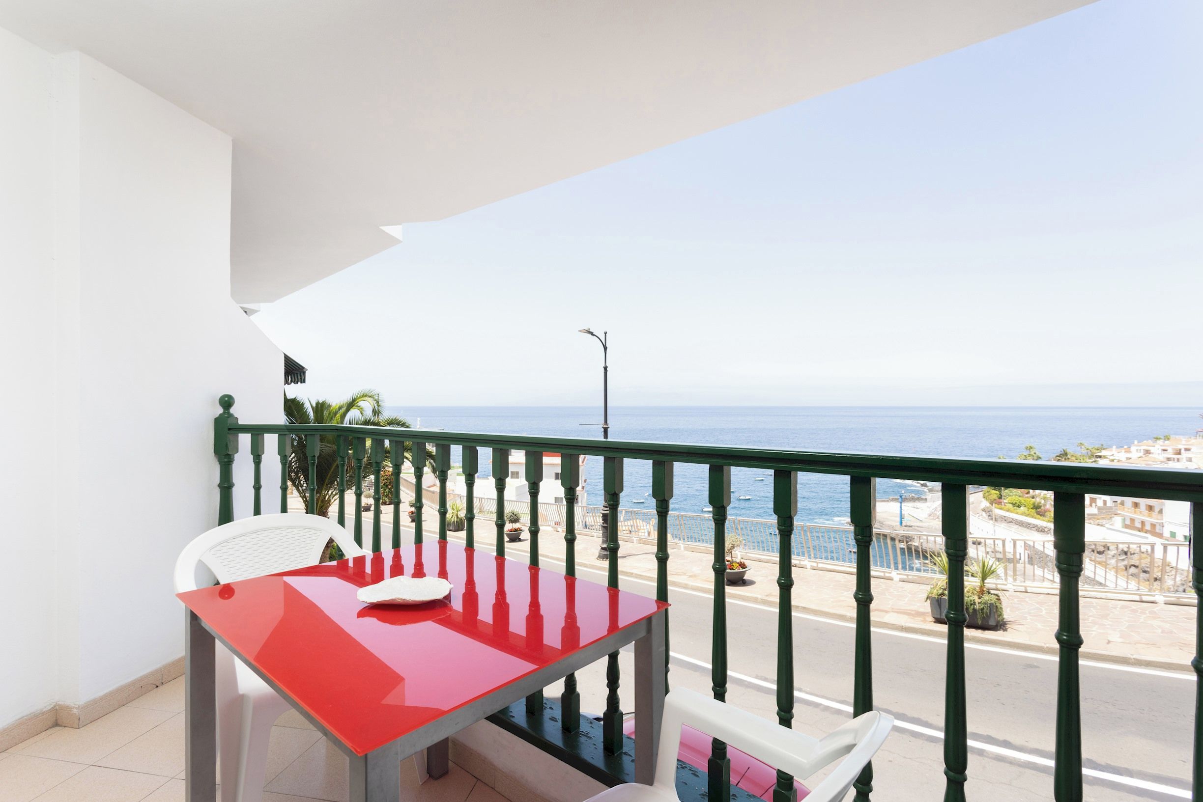 Ein Balkon mit einem roten Tisch und weißen Stühlen, der Blick auf die Küste mit Meer und Gebäuden freigibt.