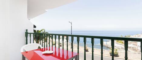 A balcony with a red table and white chairs, overlooking a coastal view with the sea and buildings.