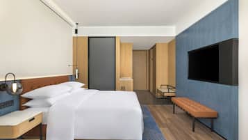 A modern hotel room with a bed, bedside table, lamp, bench, TV, and a door leading to another room.