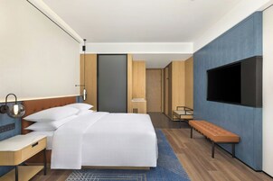 Habitación de hotel moderna con cama, mesita de noche, lámpara, banco, televisor y puerta que da a otra habitación.