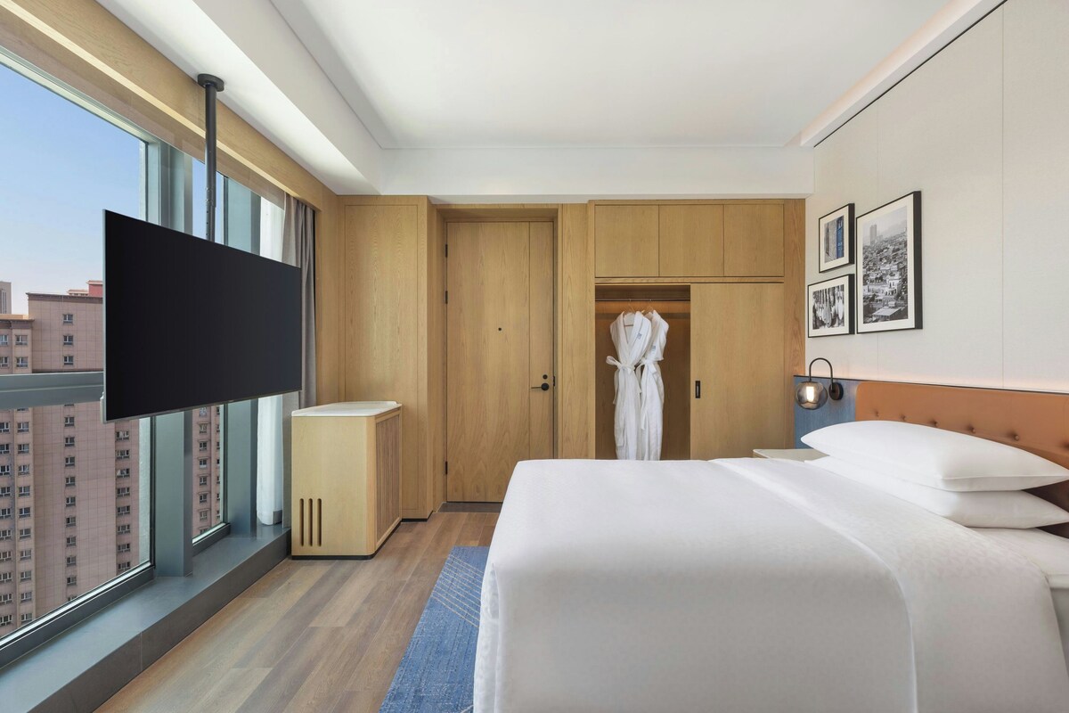 A hotel room with a large bed, a TV, a wardrobe, and a view of buildings outside.
