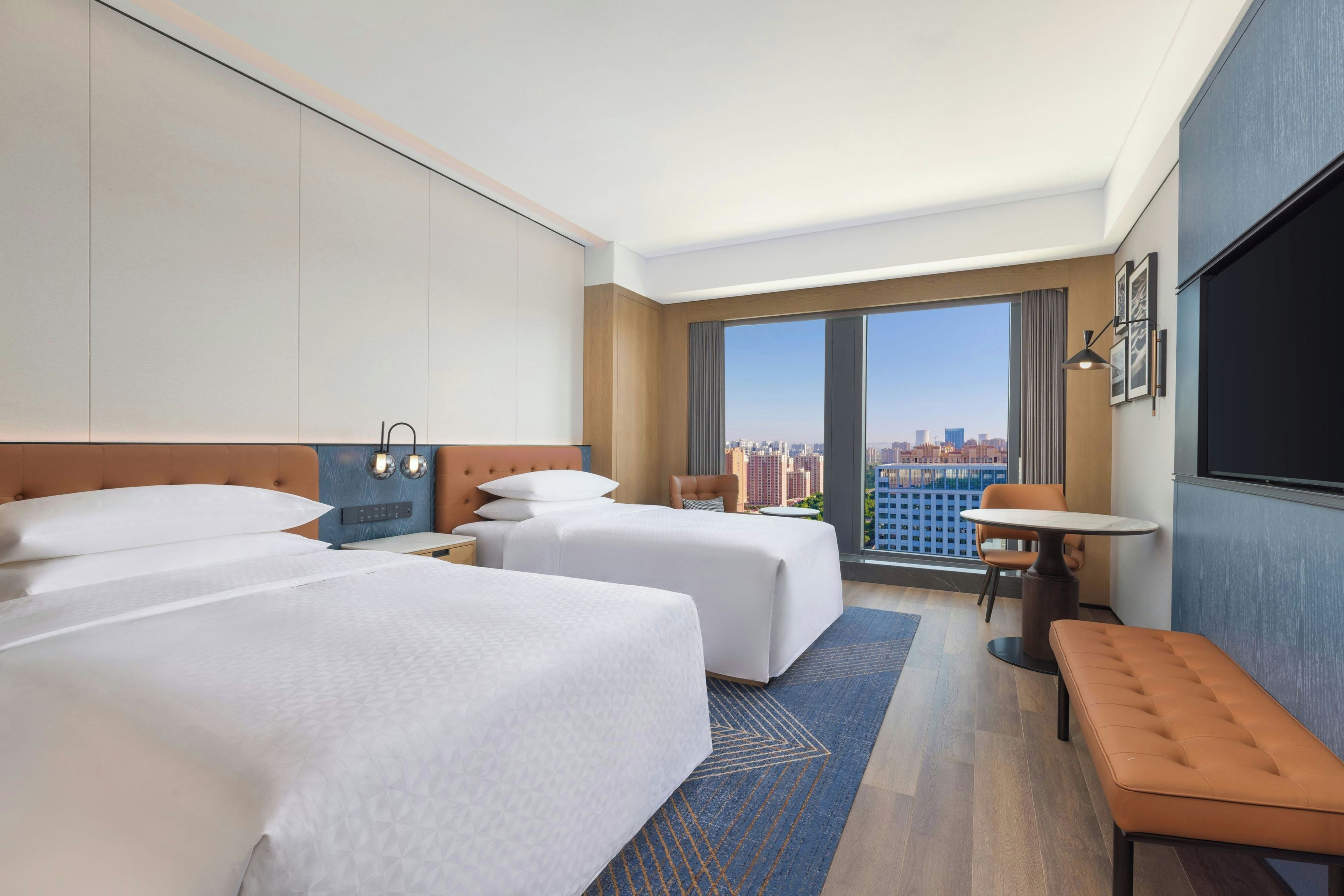 A hotel room with two beds, a small table, a bench, a TV, and a large window offering a city view.