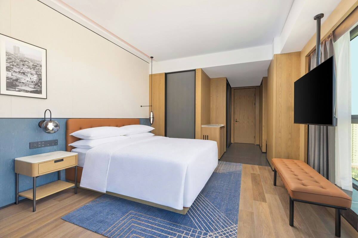 A hotel room with a bed, bedside table, bench, TV, and a painting on the wall.