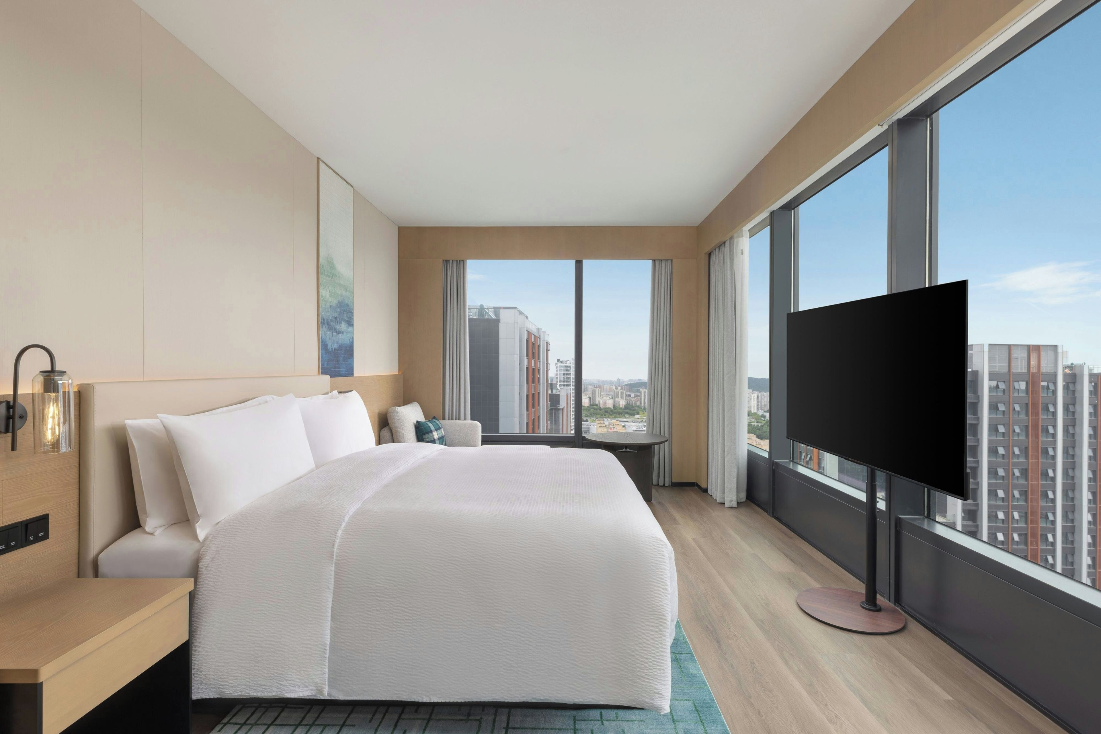 A hotel room with a large bed, a TV, a desk, and a view of the city.