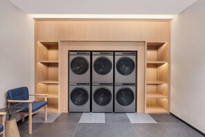 A laundry room with six front-loading washing machines, wooden shelving, a blue chair, and a small wooden table. - Fairfield By Marriott Nanjing Yuhuatai (Nanjing)