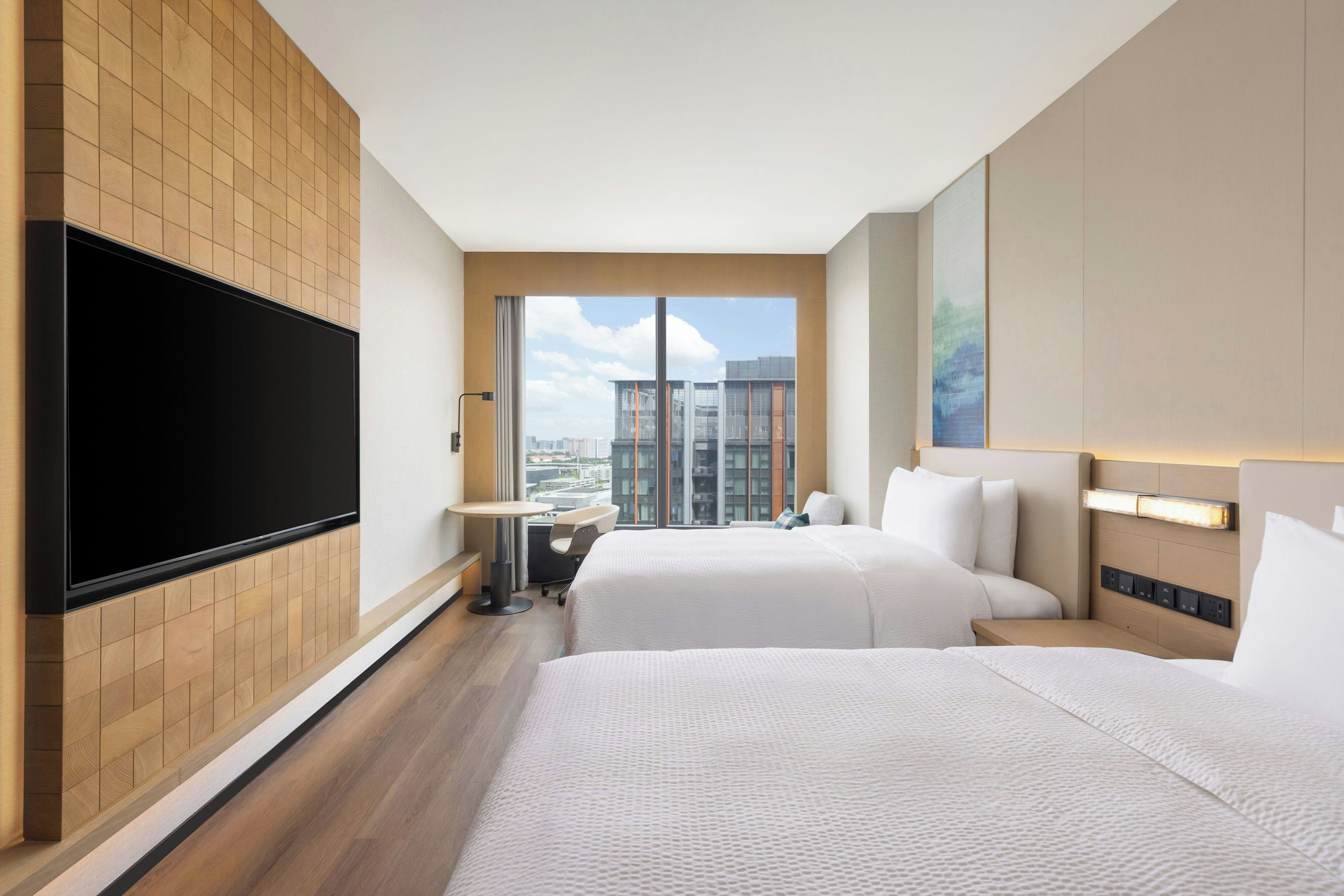 A hotel room with two beds, a flat-screen TV mounted on the wall, a small table, and a view of the cityscape through large windows.