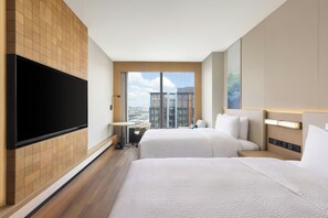 Habitación de hotel con dos camas, un televisor de pantalla plana montado en la pared, una mesita pequeña y vistas panorámicas de la ciudad a través de amplios ventanales.