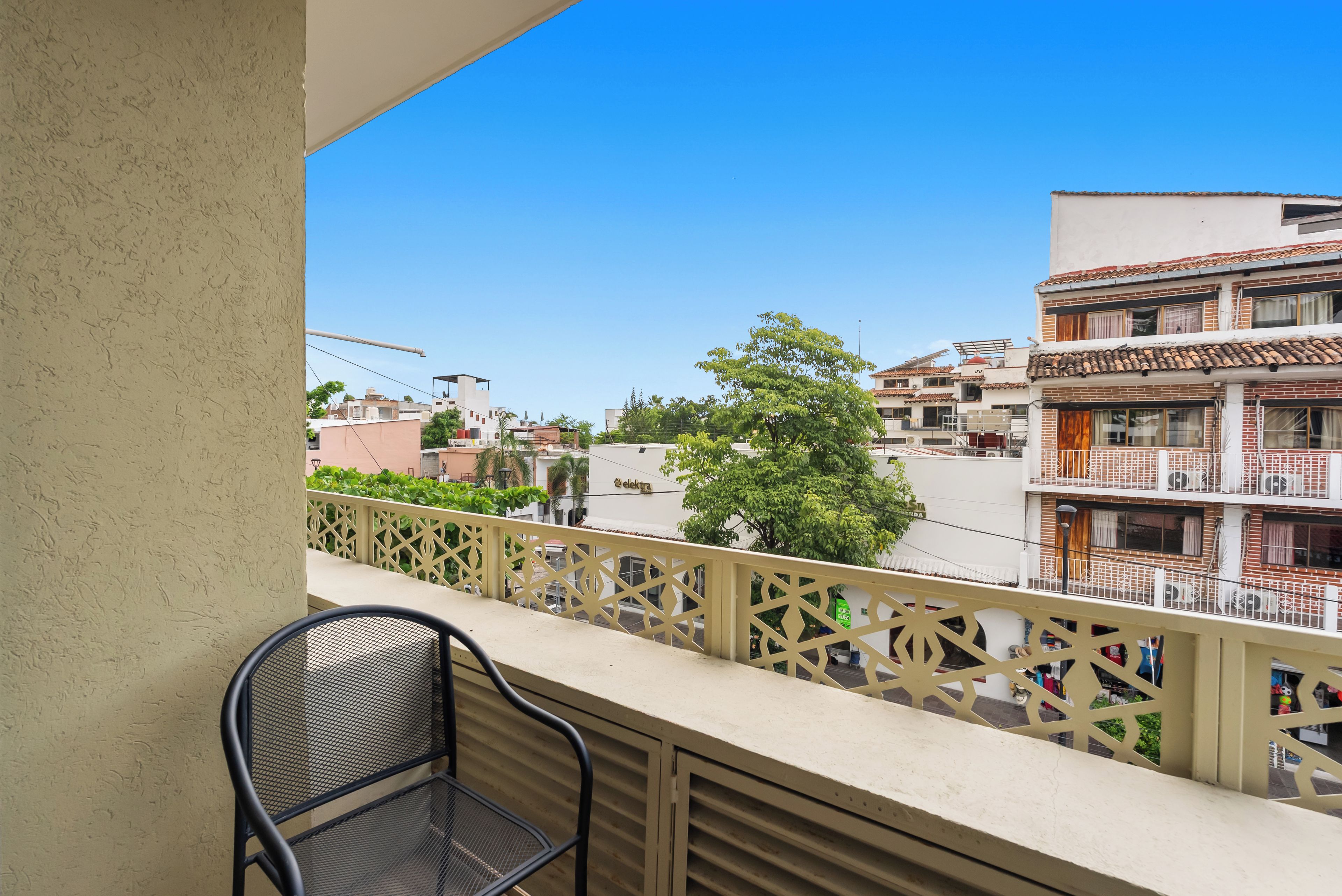 Un balcon avec une chaise en métal, une balustrade décorative et une vue sur les bâtiments voisins et un arbre.