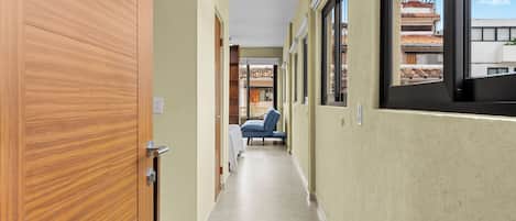 A corridor with a wooden door, a bed with a blue cover, and a window with a view of buildings.