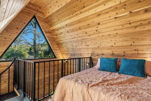 A bedroom with a wooden ceiling, a bed with patterned bedding, a window, and a balcony railing.