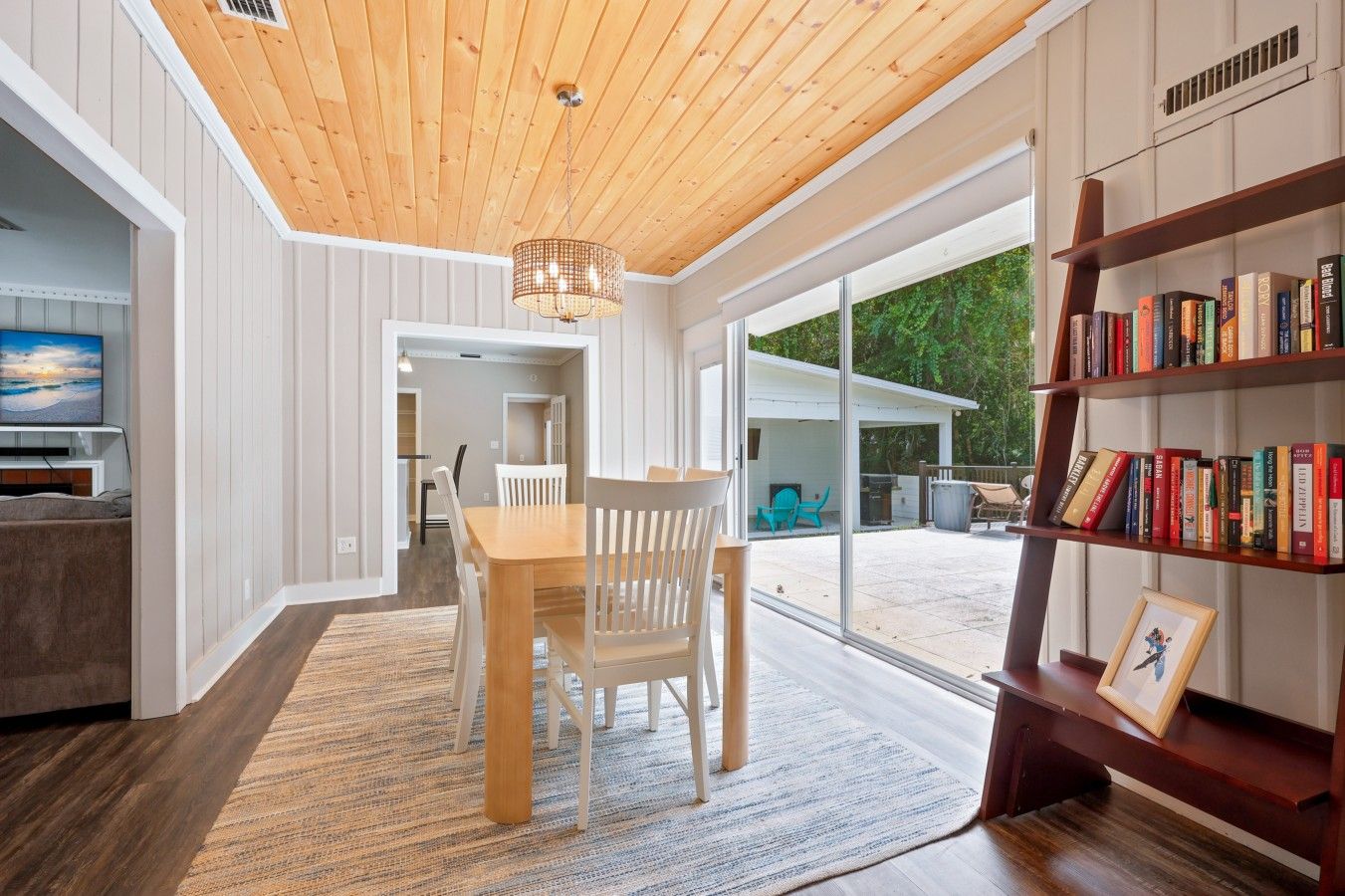 A modern interior with a wooden ceiling, a dining area with a table and chairs, a bookshelf, and a view through large glass doors to an outdoor patio.