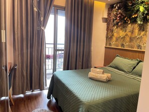 A hotel room with a bed, a window with curtains, a wall with a leaf pattern, and a wooden floor. - Modern studio in the city center | Pool + Gym | Subway (São Paulo)