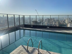 An indoor swimming pool with clear glass walls, a tiled floor, and metal pool ladders. - Modern studio in the city center | Pool + Gym | Subway (São Paulo)