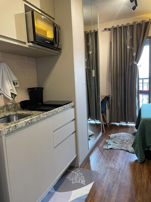 A compact kitchen with a microwave, a sink, and a coffee maker. There is a small dining area with a chair and a table. - Modern studio in the city center | Pool + Gym | Subway (São Paulo)