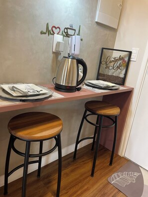 A kitchenette with a stainless steel coffee maker, two bar stools, a framed picture on the wall, and a small rug on the floor. - Modern studio in the city center | Pool + Gym | Subway (São Paulo)