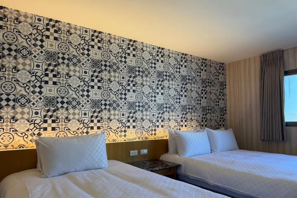 Habitació d'hotel amb dos llits, un mur amb un disseny i una finestra amb cortines.