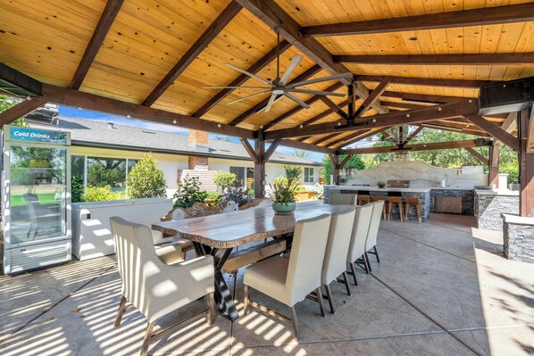 An outdoor covered patio with a dining table and chairs, a grill station, and a cold drink vending machine.