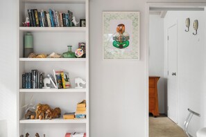 A bookshelf with various books and decorative items, a framed artwork, and a door leading to another room. - Slice of Paradise - Whangamata Holiday Home (Whangamata)