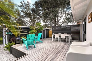 A wooden deck with outdoor seating, a barbecue grill, and a fenced garden area. - Slice of Paradise - Whangamata Holiday Home (Whangamata)