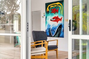 A room with a large shark-themed painting on the wall, a wooden chair, and a glass door leading outside. - Slice of Paradise - Whangamata Holiday Home (Whangamata)