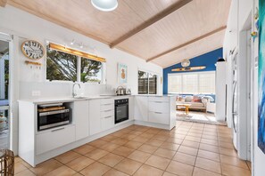 A modern kitchen with white cabinetry, a built-in oven, and a window with a view of trees. - Slice of Paradise - Whangamata Holiday Home (Whangamata)