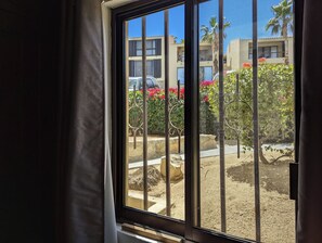 Blick durch ein Fenster mit Metallgitterstäben, im Hintergrund ein Gebäude und ein sandiger Bereich mit Pflanzen.